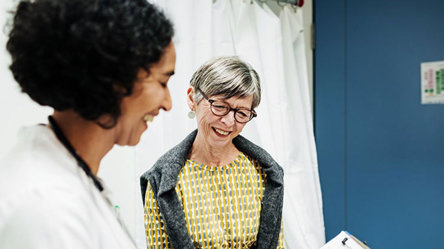 A doctor and patient in a room, the doctor is holding a clipboard with pages on it. Both people are smiling and looking at the clipboard