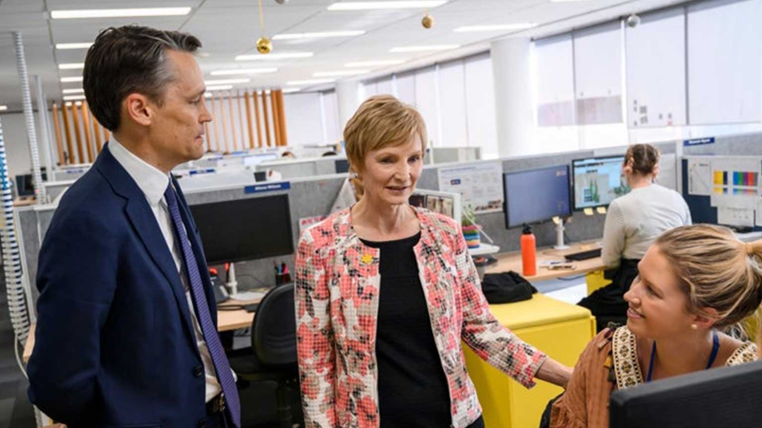 Four people working in an office, one is sitting at their computer in the background and two are standing next to a woman sitting down, speaking.