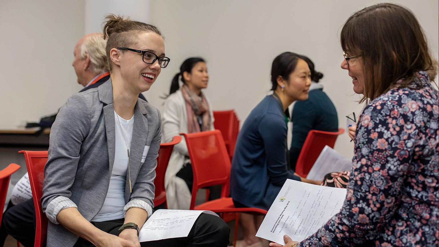 People sitting in a room, they are in pairs sitting opposite each other on red seats, talking and smiling.